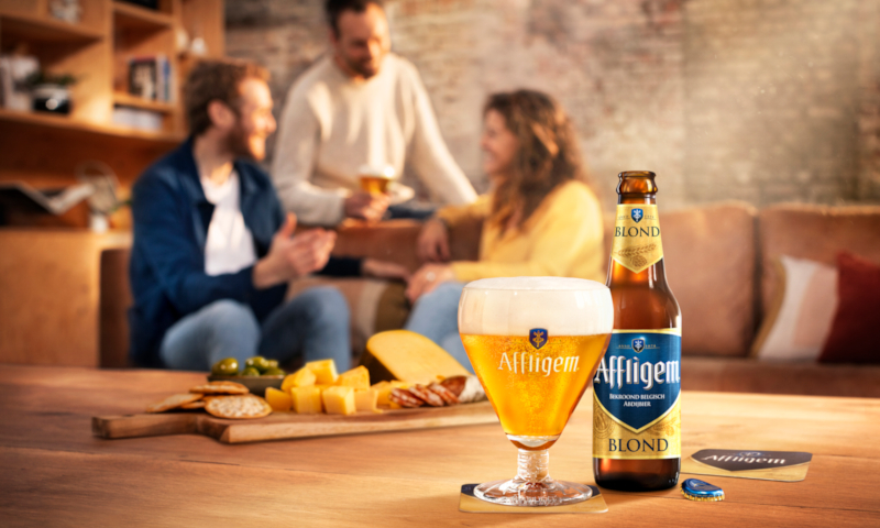 Affligem Blond glas en nieuwe retourfles op tafel met hapjes en drie mensen die erachter op de bank zitten